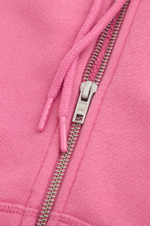 Close-up of the Dairy Boy Strawberry Milk Bottle Zip Up in strawberry pink, highlighting its metal zipper with DB engraved and two matching drawstrings with plastic tips, resting on the soft fabric.