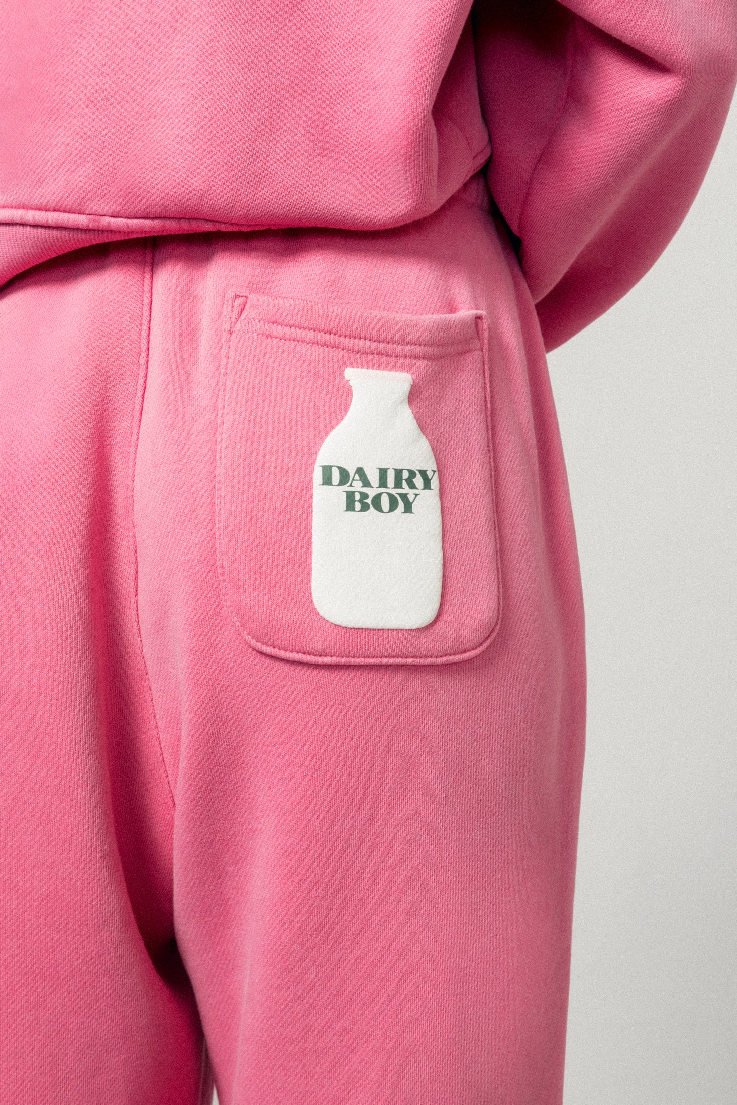 Close-up of a person in Dairy Boy's Milk Bottle Sweatpant in Strawberry Milk, featuring a back pocket with a white milk bottle print labeled DAIRY BOY. Matching pink sweatshirt is partially visible.