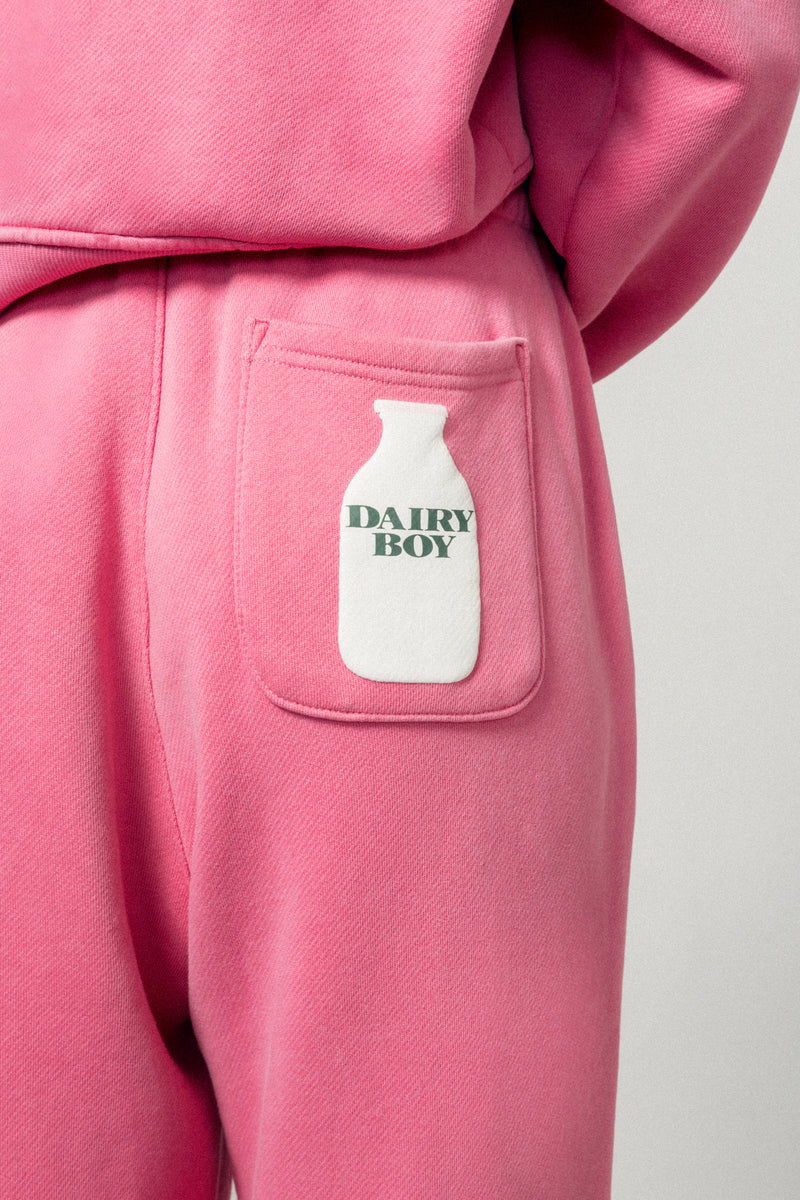 Close-up of a person in Dairy Boy's Milk Bottle Sweatpant in Strawberry Milk, featuring a back pocket with a white milk bottle print labeled DAIRY BOY. Matching pink sweatshirt is partially visible.