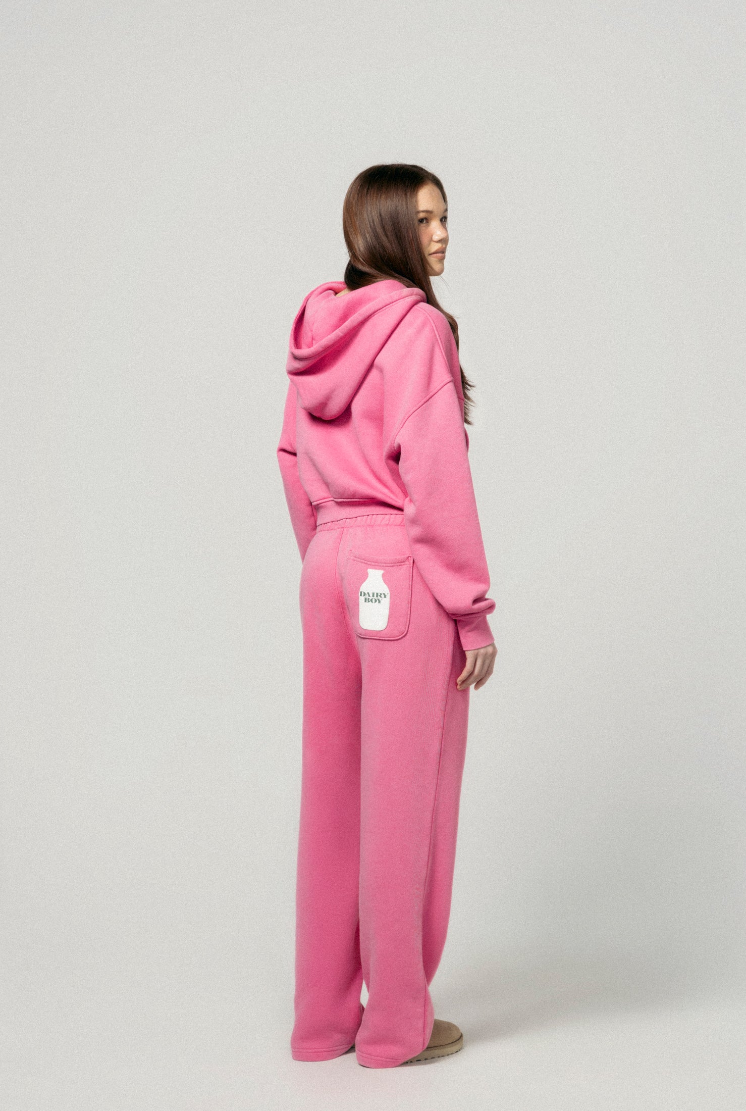 A woman faces away, wearing the Dairy Boy Strawberry Milk Bottle Zip Up and matching strawberry pink sweatpants with a white milk bottle on the right back pocket. The background is plain light gray.