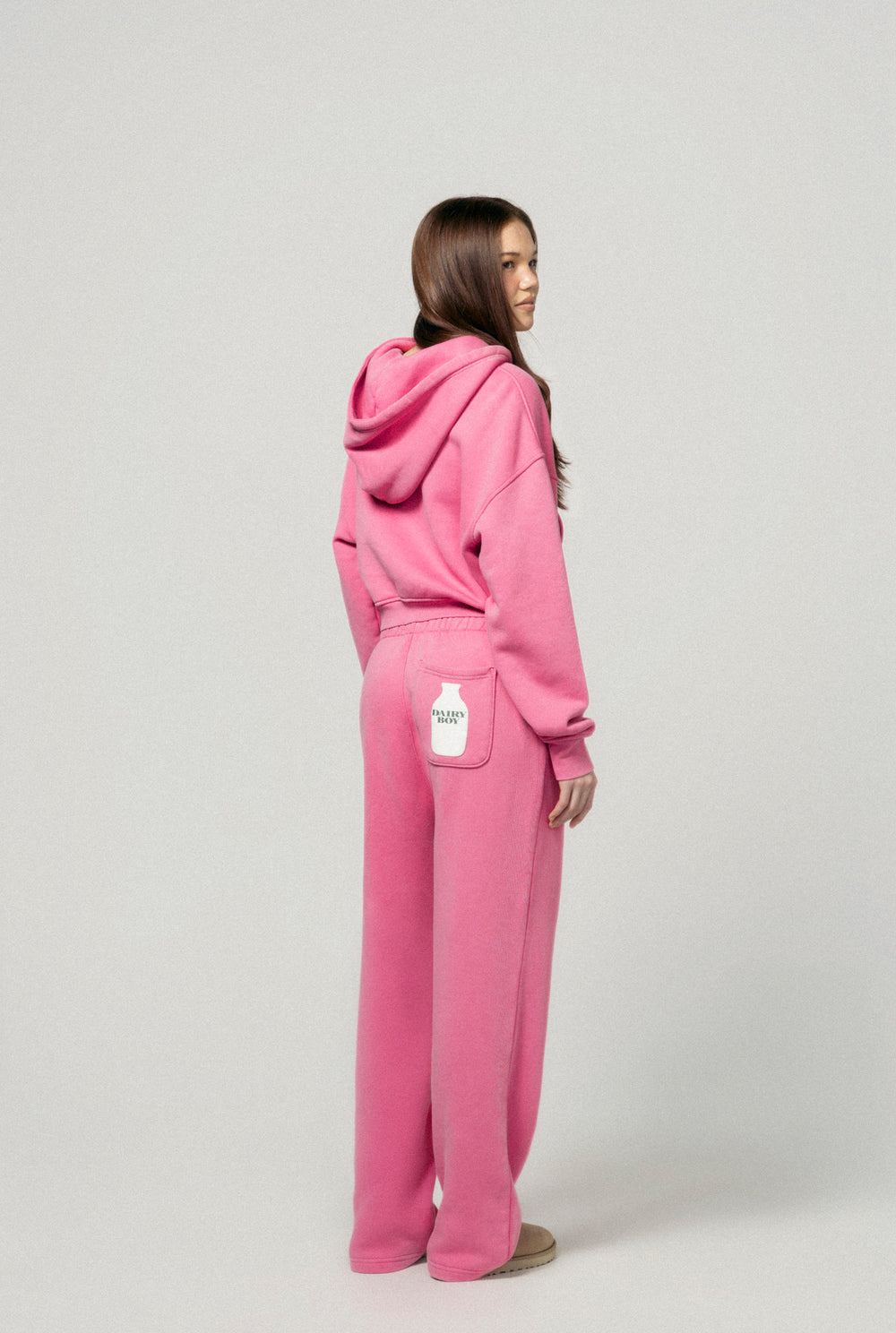 A young woman with long brown hair, wearing Dairy Boy's Milk Bottle Sweatpant in Strawberry Milk, stands facing away with a white milk bottle on the back pocket. The background is light gray.