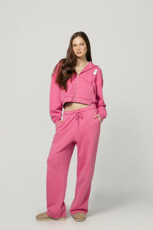 A young woman with long brown hair faces forward, wearing Dairy Boy's Strawberry Milk Milk Bottle Sweatpant and matching pink cropped hoodie, hands in pockets. She pairs the look with tan slippers against a plain light gray background.