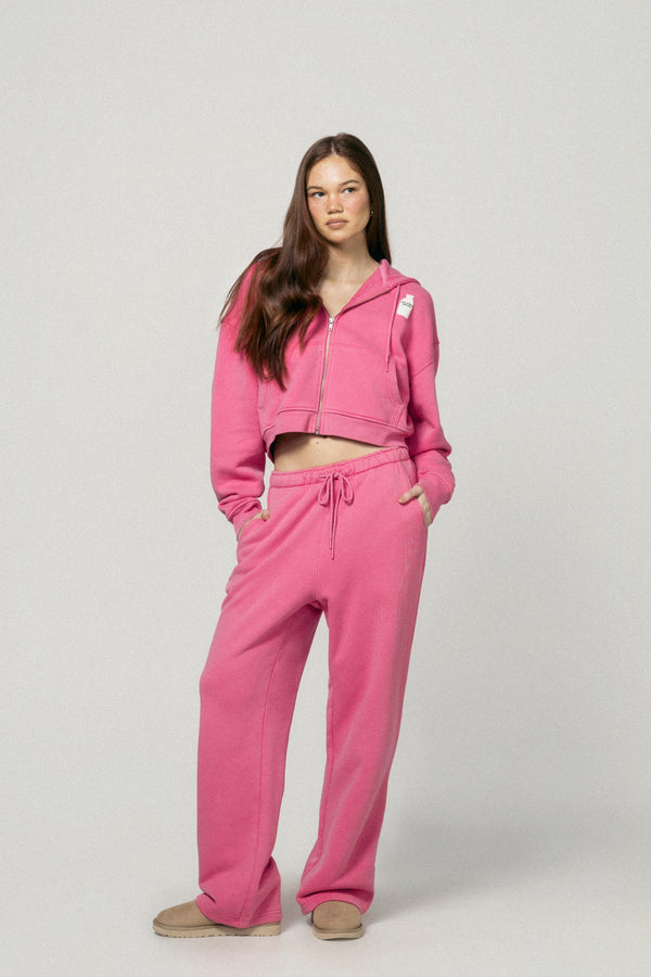 A young woman with long brown hair faces forward, wearing Dairy Boy's Strawberry Milk Milk Bottle Sweatpant and matching pink cropped hoodie, hands in pockets. She pairs the look with tan slippers against a plain light gray background.