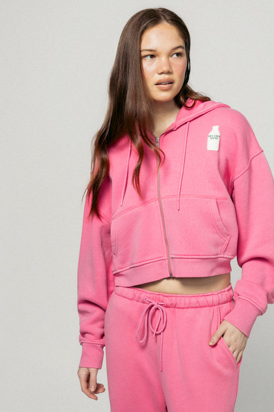 A young woman with long brown hair wears the Dairy Boy Strawberry Milk Bottle Zip Up in strawberry pink, paired with matching sweatpants. She stands against a plain light gray background, gazing off to the side with a relaxed expression. |m1