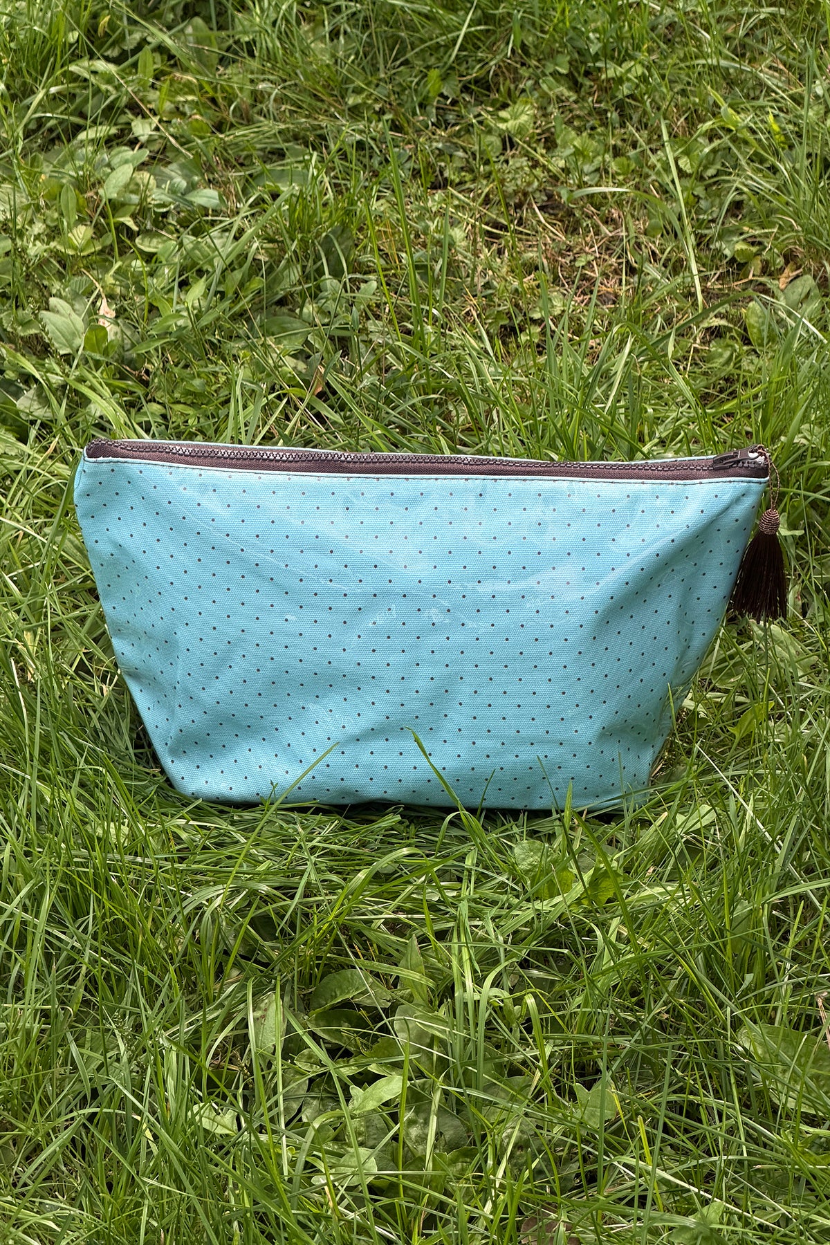 Light blue makeup bag with small dark dots, a brown zip across the top and a brown tassel on the pull, front view.