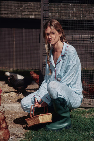 Light blue polka-dot pajama set with burgundy piping and a chest pocket, worn on a model crouching with a wicker basket. |M2
