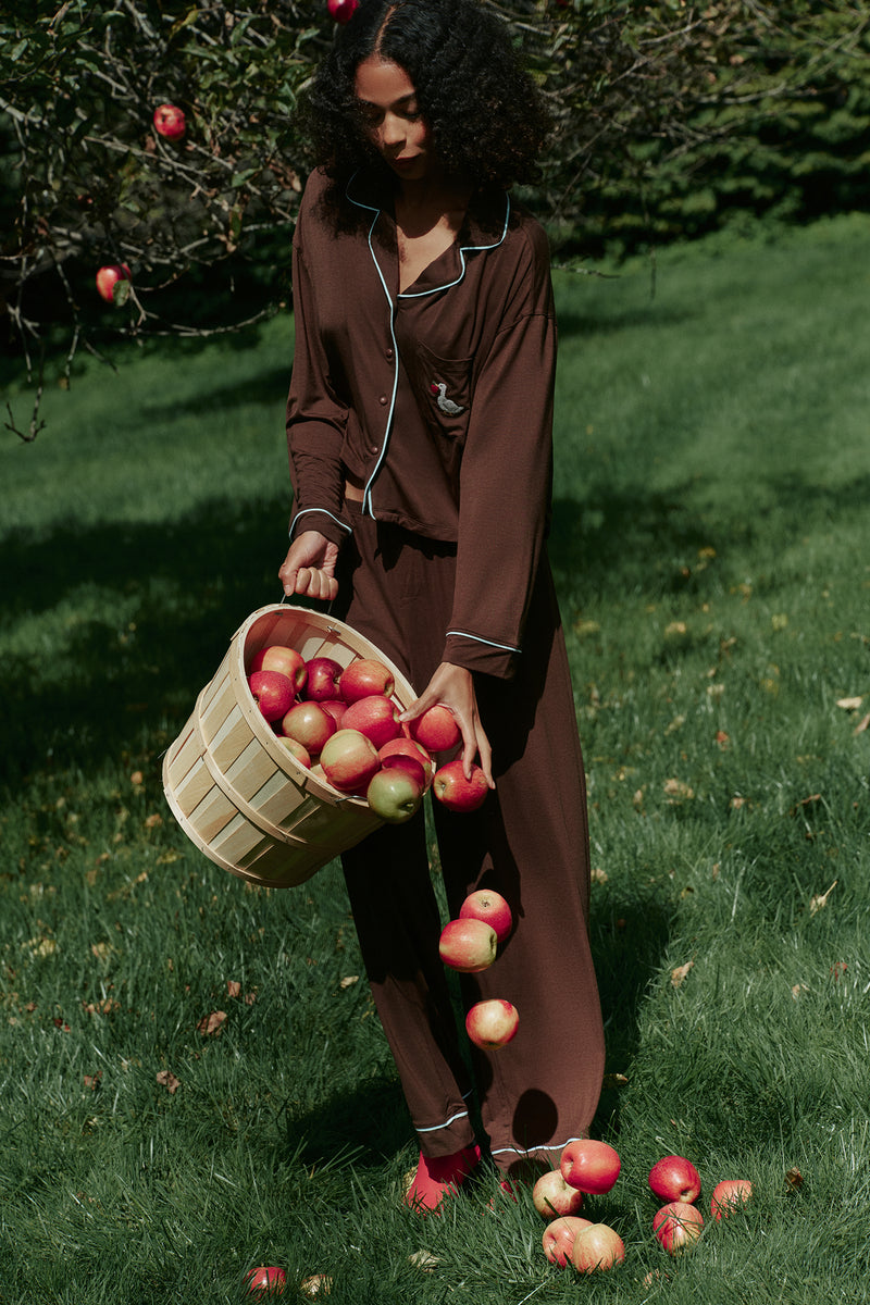 Brown long-sleeve pajama set with light blue piping, buttons and a small chest-pocket motif, worn on a model with wide-leg pants. |M1