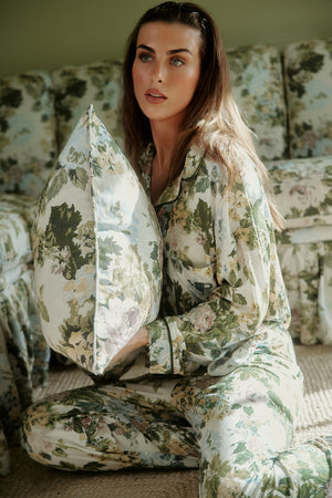 Long-sleeve pajama set in a green, cream and pale blue floral print with dark piping and a chest pocket, worn on a model holding a matching cushion. |M1
