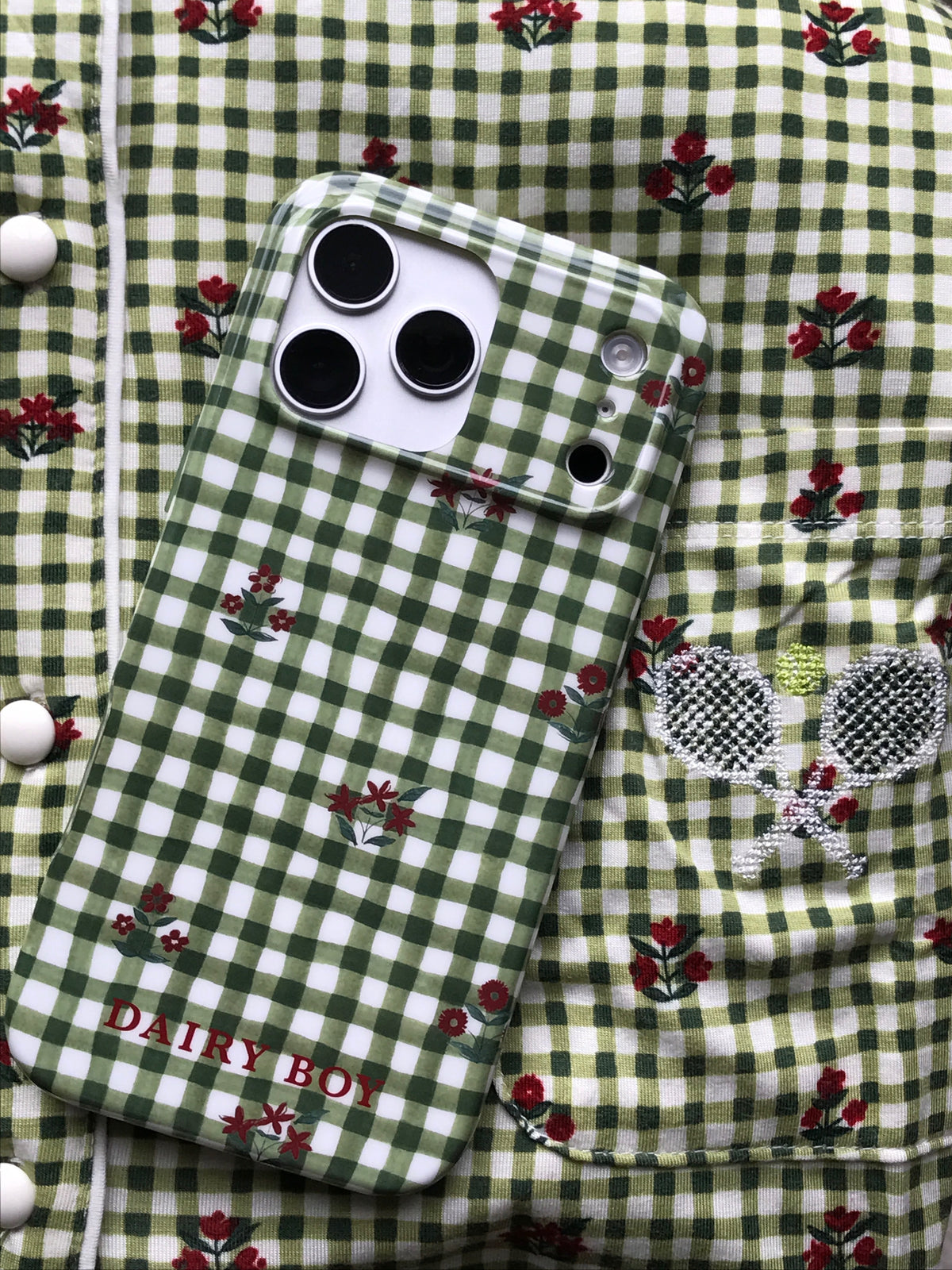 Green and white gingham phone case with small red floral sprigs and red "DAIRY BOY" lettering, back view resting on matching fabric.